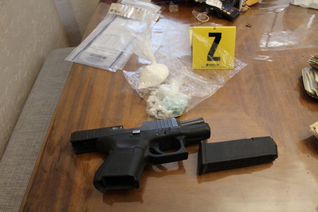 A black handgun with its detached magazine sits on a wooden table alongside several small plastic bags containing white substances and blue bills